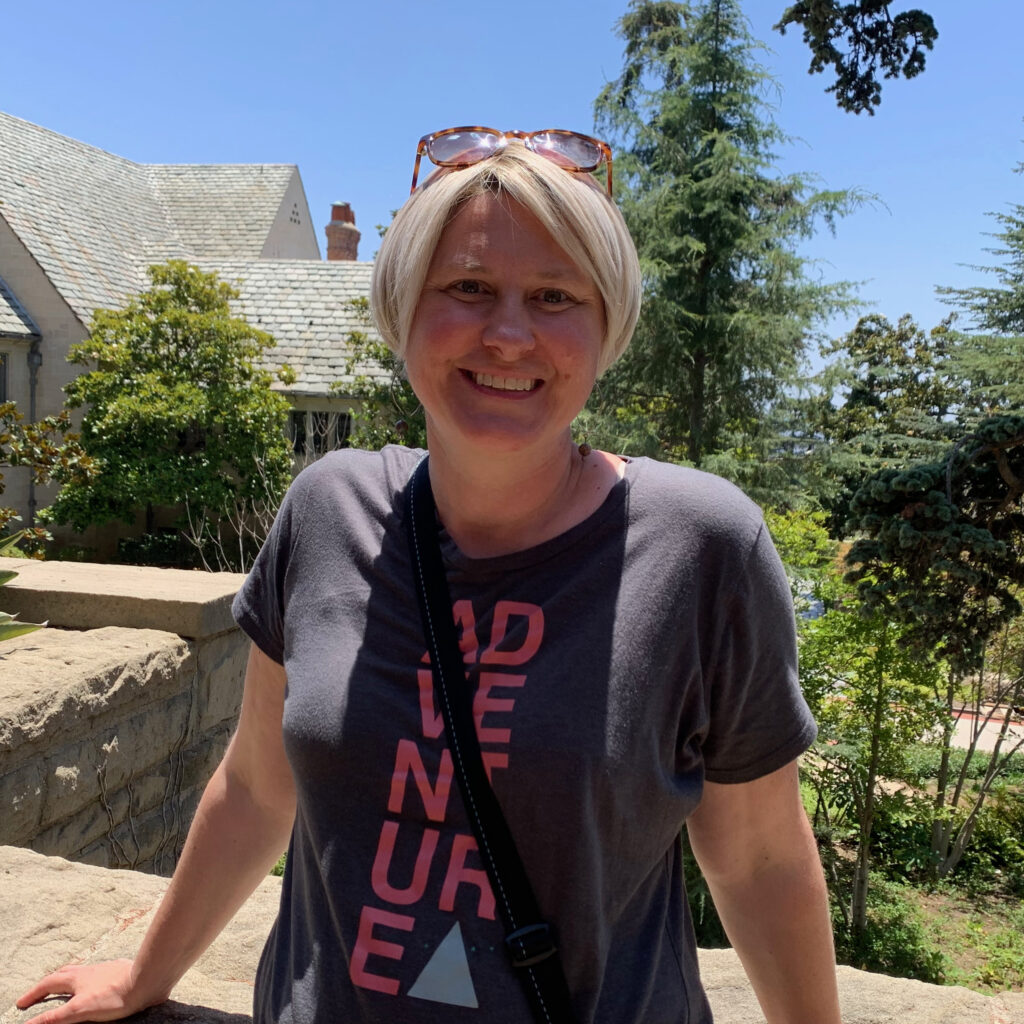 Karis, a white woman with short blond hair and sunglasses on her head, stands in front of house in the sun, smiling.