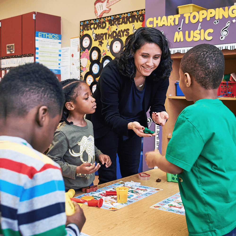 Three children gather around a teacher in a classroom setting.